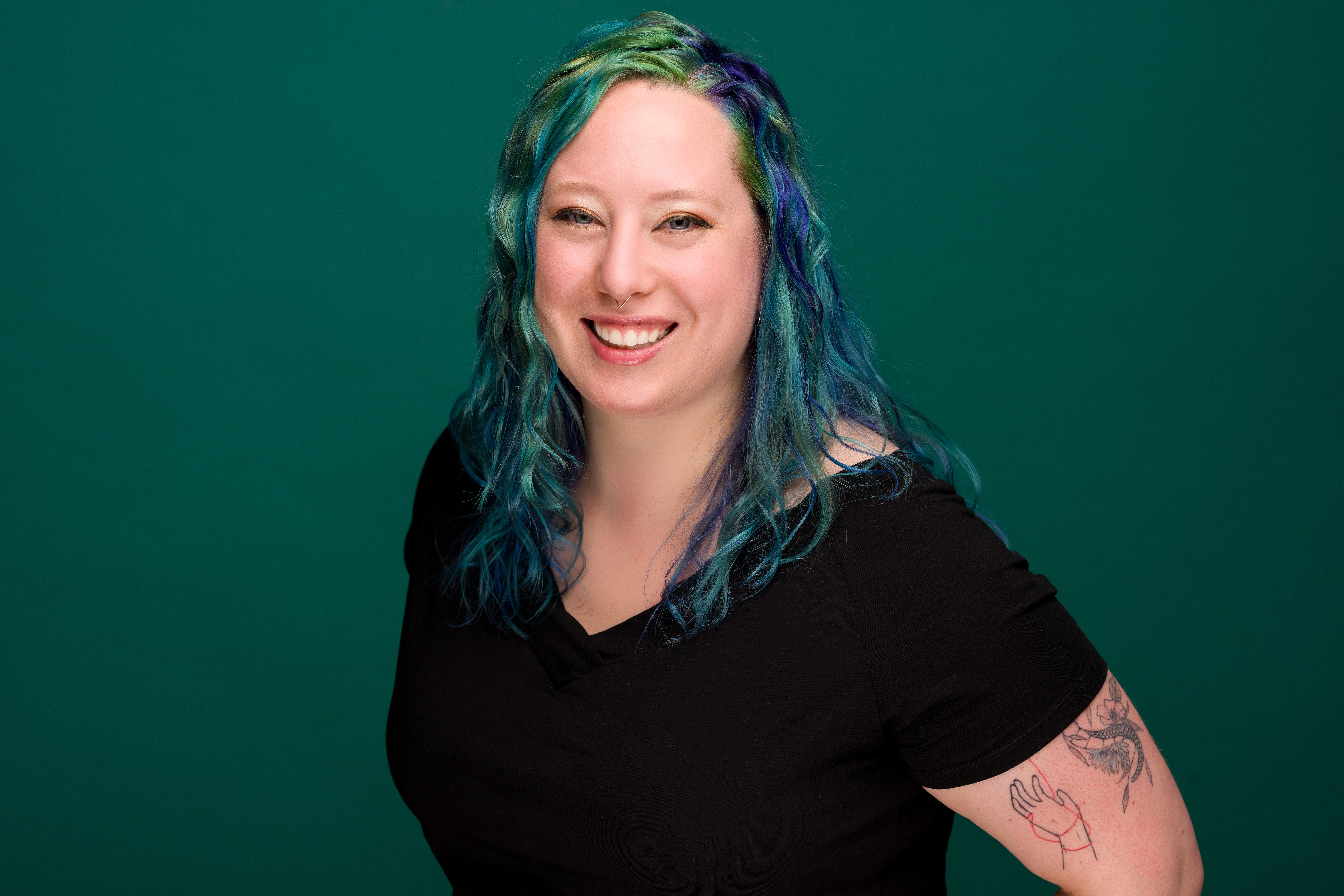 image of kat, a woman with blue and green hair, wearing a black shirt. she is angled toward the camera and smiling.