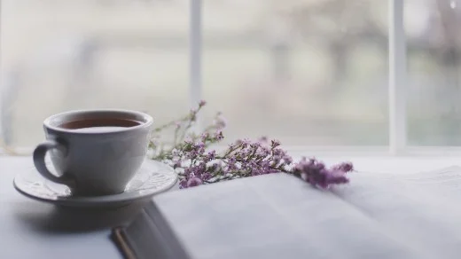 Ceramic mug of tea or coffee with flowers and a book in front of a window