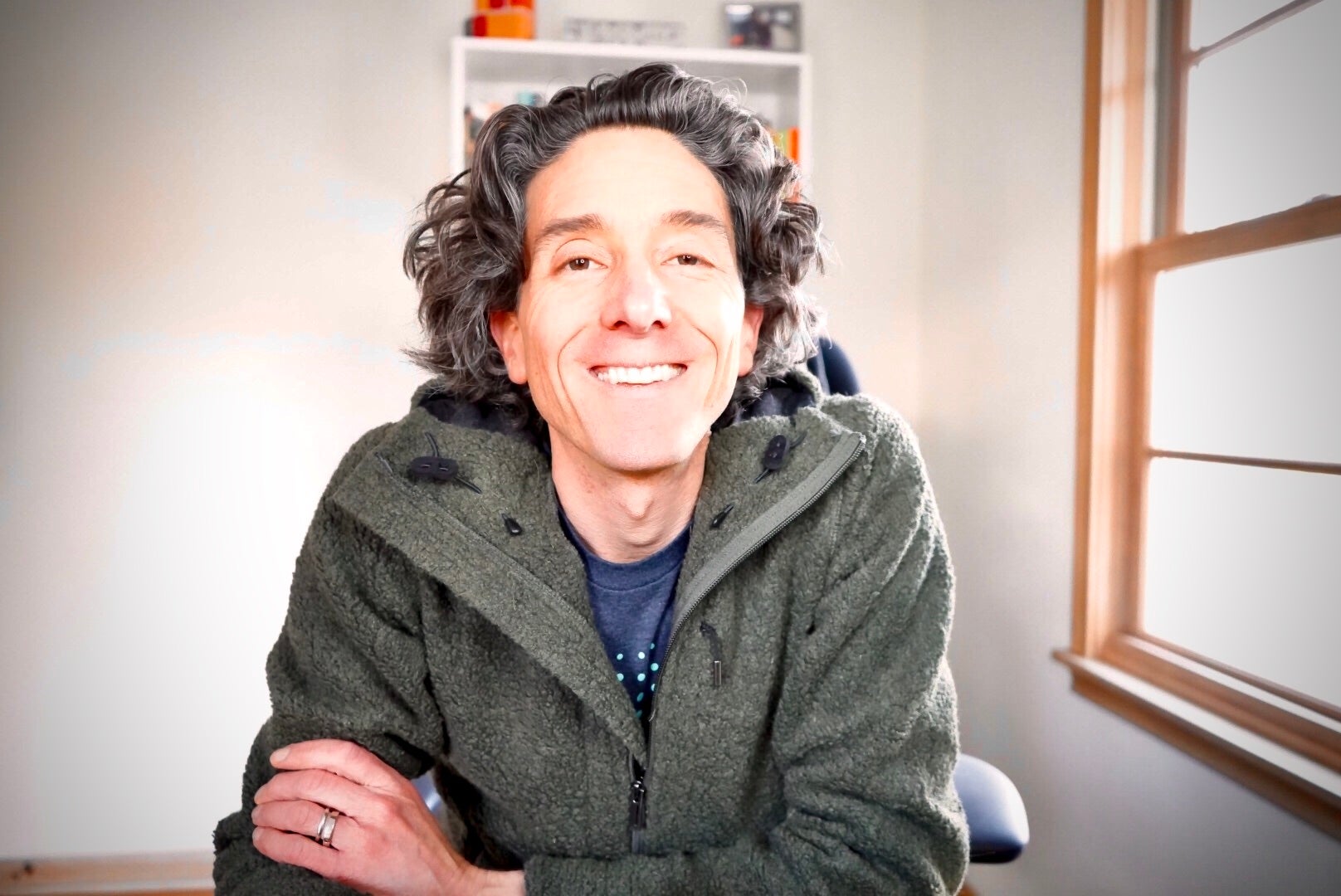 Picture of Sam in his home office smiling at the camera. Sam is a white man. He has long curly brown hair and is wearing a dark green zip up fleece hoody.