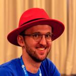 A close-up portrait of a smiling white man with short dark hair and a neatly trimmed beard. He is wearing a red fedora hat, a blue shirt, and a blue lanyard with the word "Fedora" printed on it. The background is blurred, suggesting he is indoors.