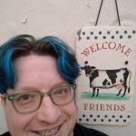 Blue-haired Eric next to a slate shingle with a cow and the word "welcome" painted upon it.