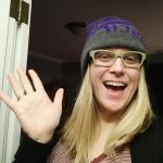A blonde woman who is waving as though saying "Hi!", wearing a gray and purple snow hat and beige glasses