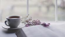 Ceramic mug of tea or coffee with flowers and a book in front of a window
